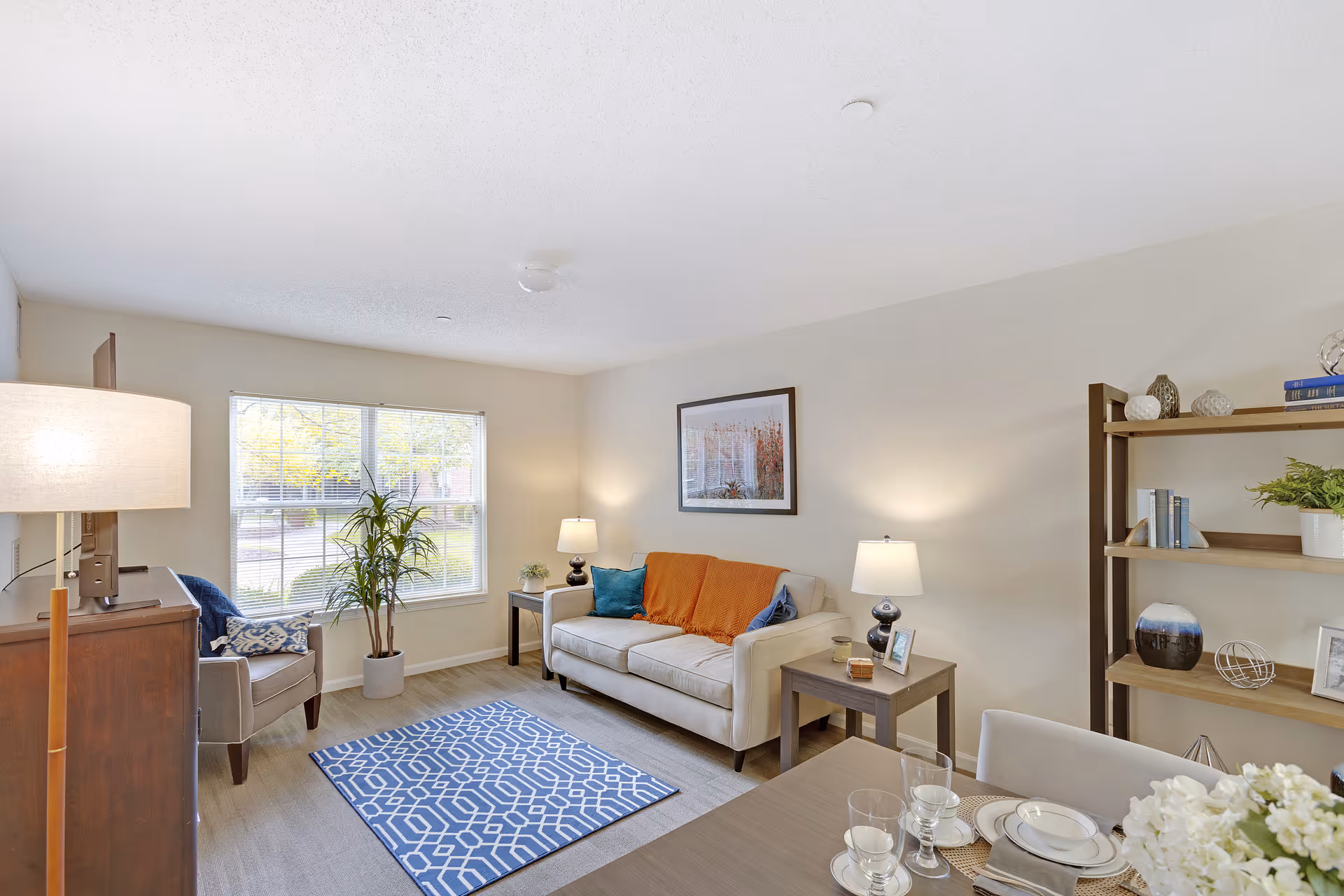 A bright and cozy living room with a beige sofa adorned with blue and orange pillows, two side tables with lamps, a wooden bookshelf with decorative items, a blue patterned rug, a potted plant near a window with blinds, and a dining table set with plates and glasses in the foreground.