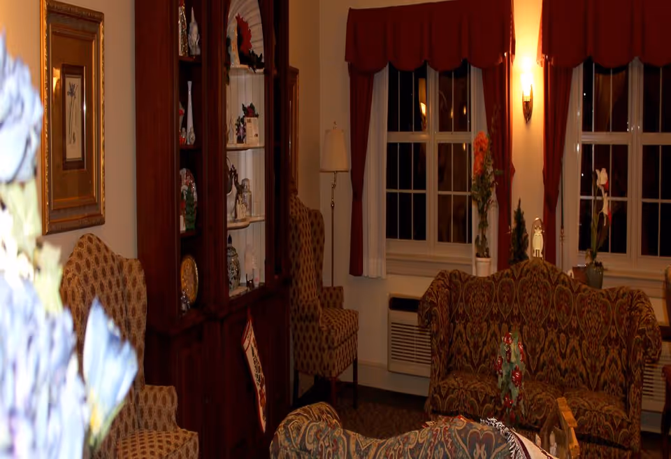 A cozy living room with patterned upholstered armchairs and a sofa, a wooden cabinet displaying decorative items, framed artwork on the wall, a floor lamp, and windows with red curtains and white sheers. There are also small plants and holiday decorations placed around the room.