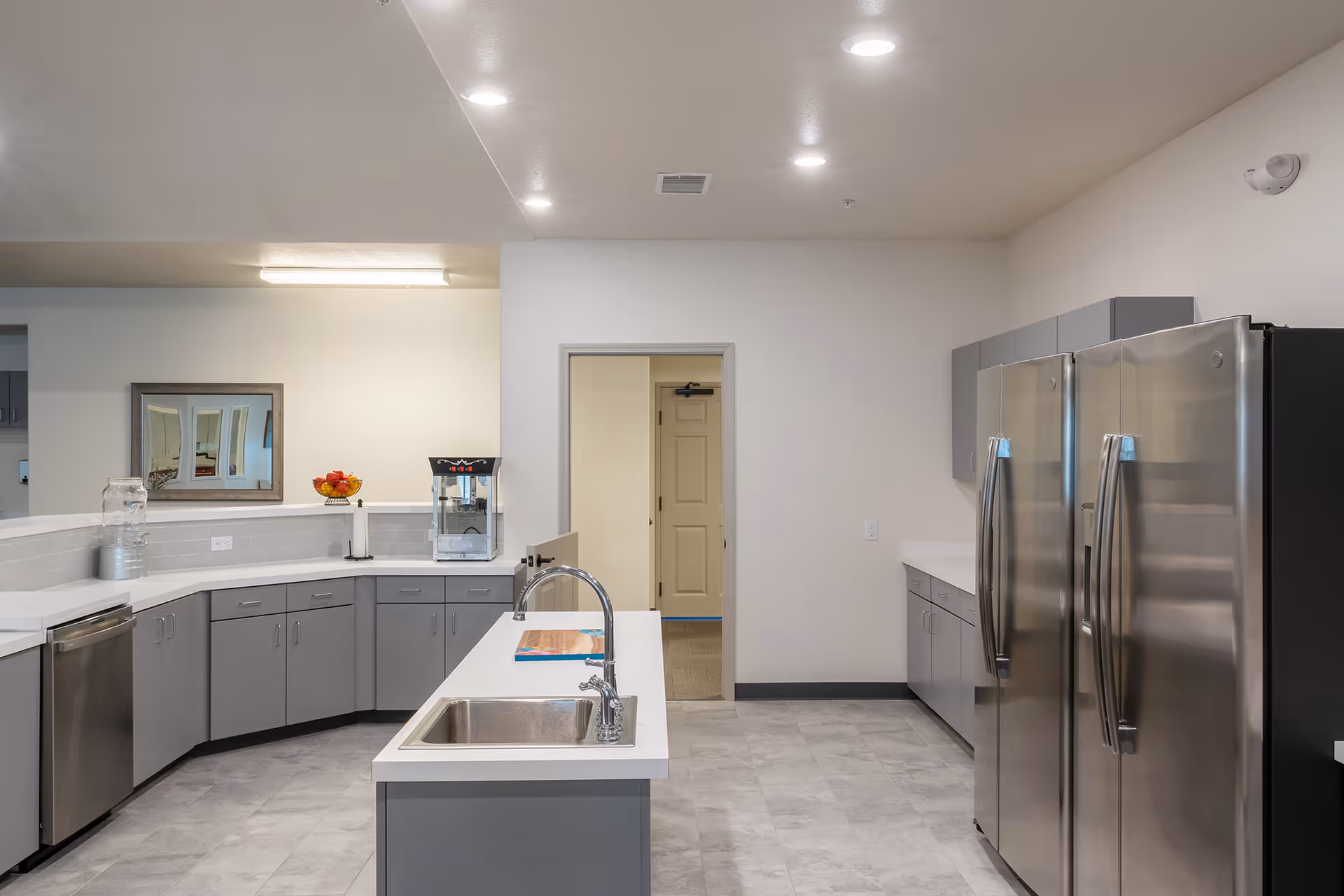 Modern kitchen area with gray cabinets, white countertops, a double-door stainless steel refrigerator, a dishwasher, a sink on an island counter, a popcorn machine, and a bowl of fruit on the counter. The room has recessed lighting and a door leading to another area.