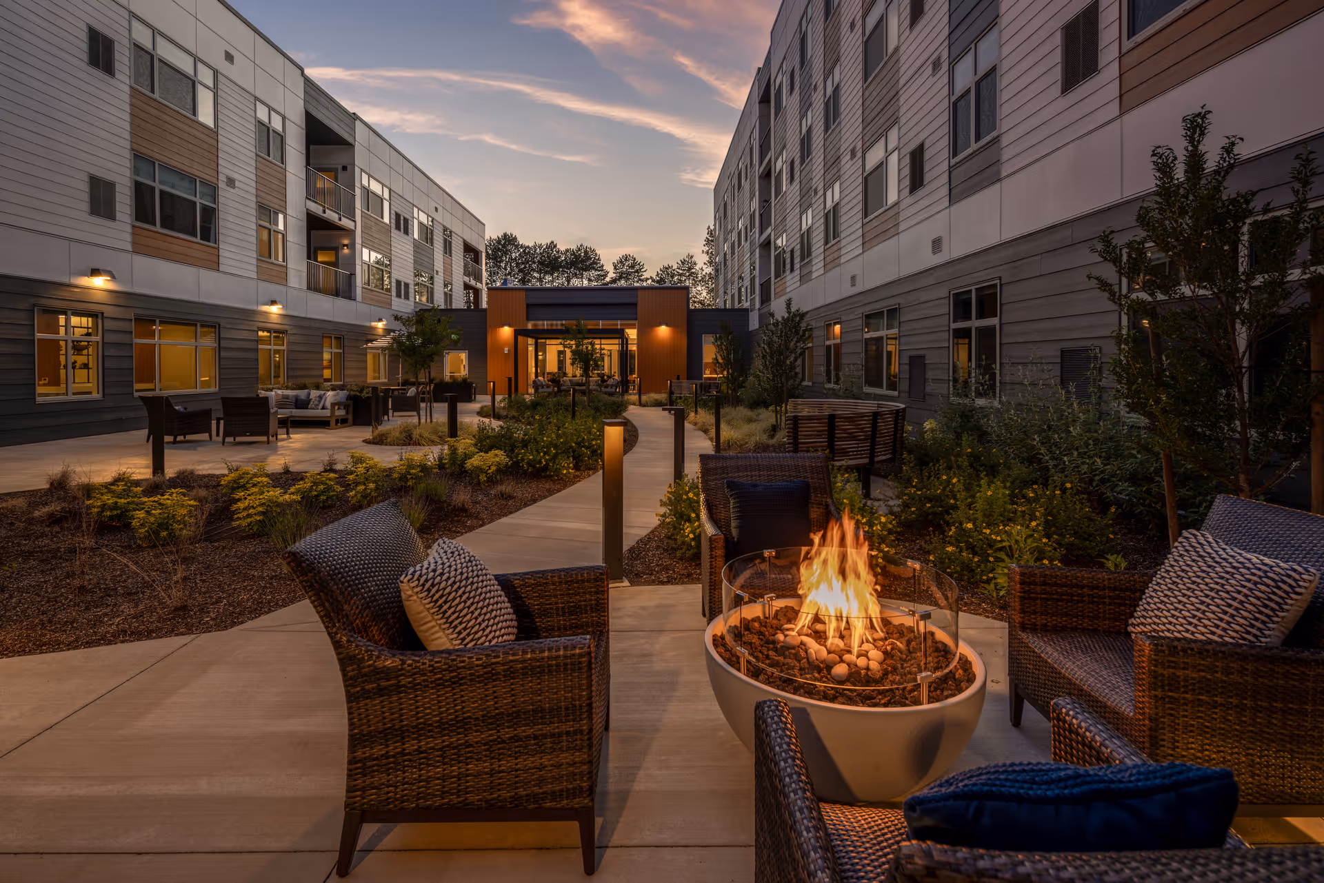 Outdoor courtyard area at sunset with modern wicker chairs arranged around a circular fire pit. The courtyard is surrounded by a multi-story building with large windows and landscaped garden beds with shrubs and small trees. A paved walkway leads to a building entrance with warm lighting.