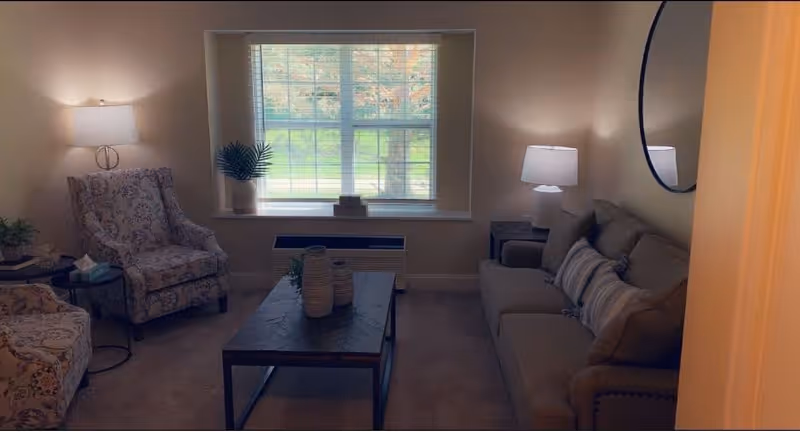 A cozy living room with a large window letting in natural light. The room features a gray sofa with striped pillows, two patterned armchairs, a dark wooden coffee table with decorative vases, two side tables with lamps, and a round mirror on the wall.