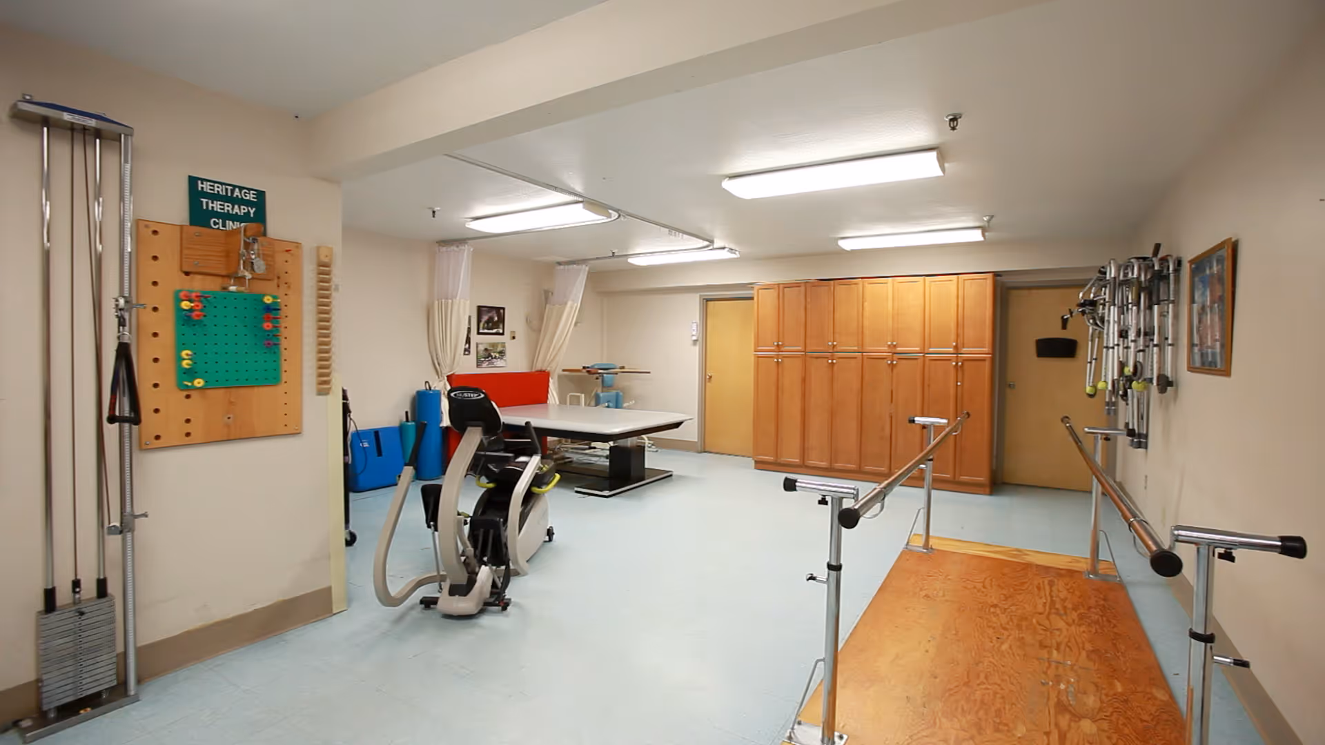 A therapy clinic room with exercise and rehabilitation equipment including parallel bars, a stationary exercise bike, a treatment table, and various therapy tools mounted on the walls. The room has light blue flooring, beige walls, wooden cabinets, and fluorescent ceiling lights.
