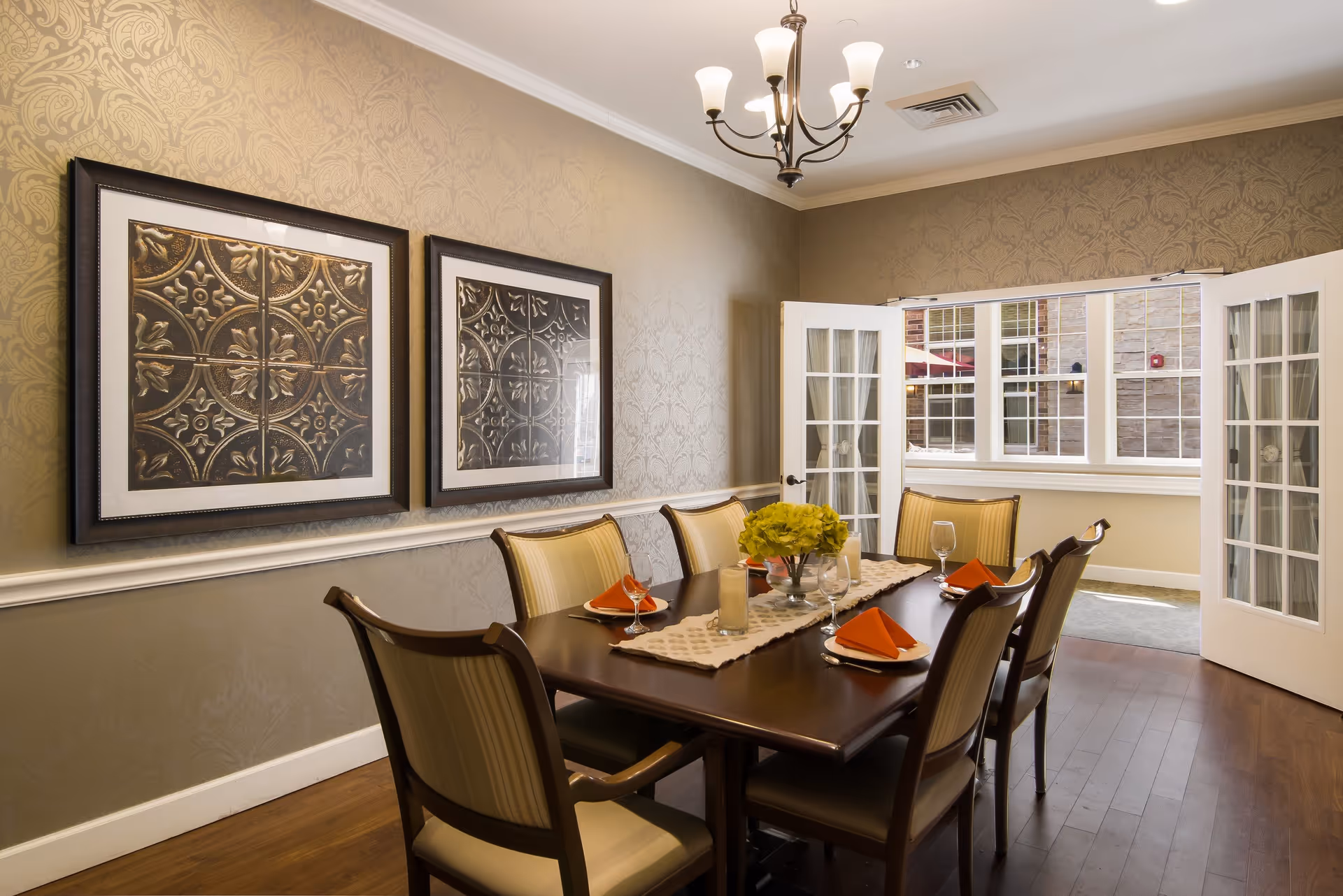 A dining room with a dark wooden table set for six people, featuring orange folded napkins, wine glasses, and a centerpiece of green flowers. The room has beige patterned wallpaper, two framed decorative art pieces on the wall, a chandelier overhead, and French doors leading to a sunlit area with large windows.