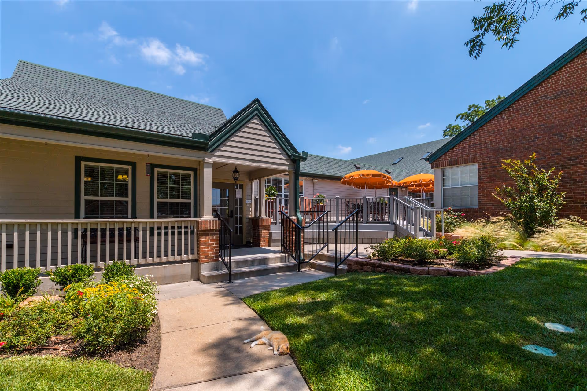 Outdoor view of Bethesda Gardens Arlington Assisted Living showing a building entrance with a small porch, steps, and railings. There are flower beds with colorful flowers and green shrubs along a concrete pathway. Two orange umbrellas shade a patio seating area. A cat is lying on the pathway in the foreground under the sunlight. The sky is clear with a few clouds.