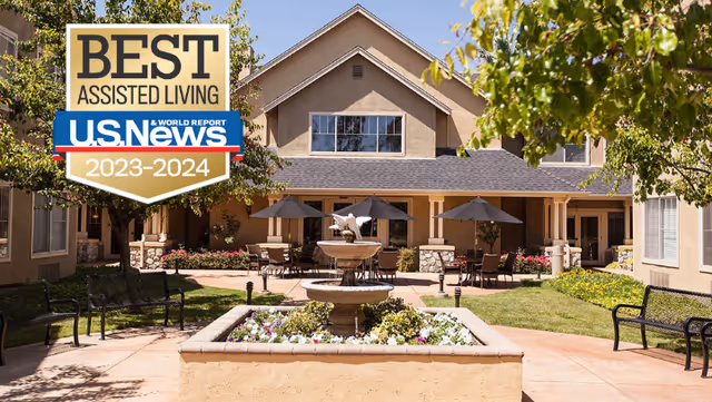 Outdoor courtyard area of Claremont Place featuring a central water fountain surrounded by flowers, patio tables with umbrellas, benches, and a two-story building in the background under a clear sky.
