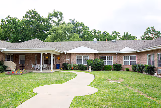 Single-story brick building with a curved concrete walkway, lawn, covered patio with chairs, and surrounding shrubs and trees.