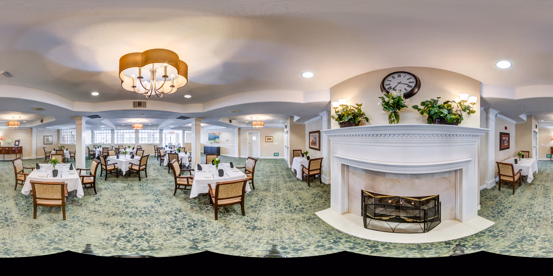 A spacious dining room with multiple tables covered in white tablecloths, each set with plates, glasses, and small flower arrangements. The room features a green patterned carpet, a large white fireplace adorned with plants and a clock above it, and several chandeliers hanging from the ceiling. The walls are light-colored with framed artwork and windows allowing natural light to enter.