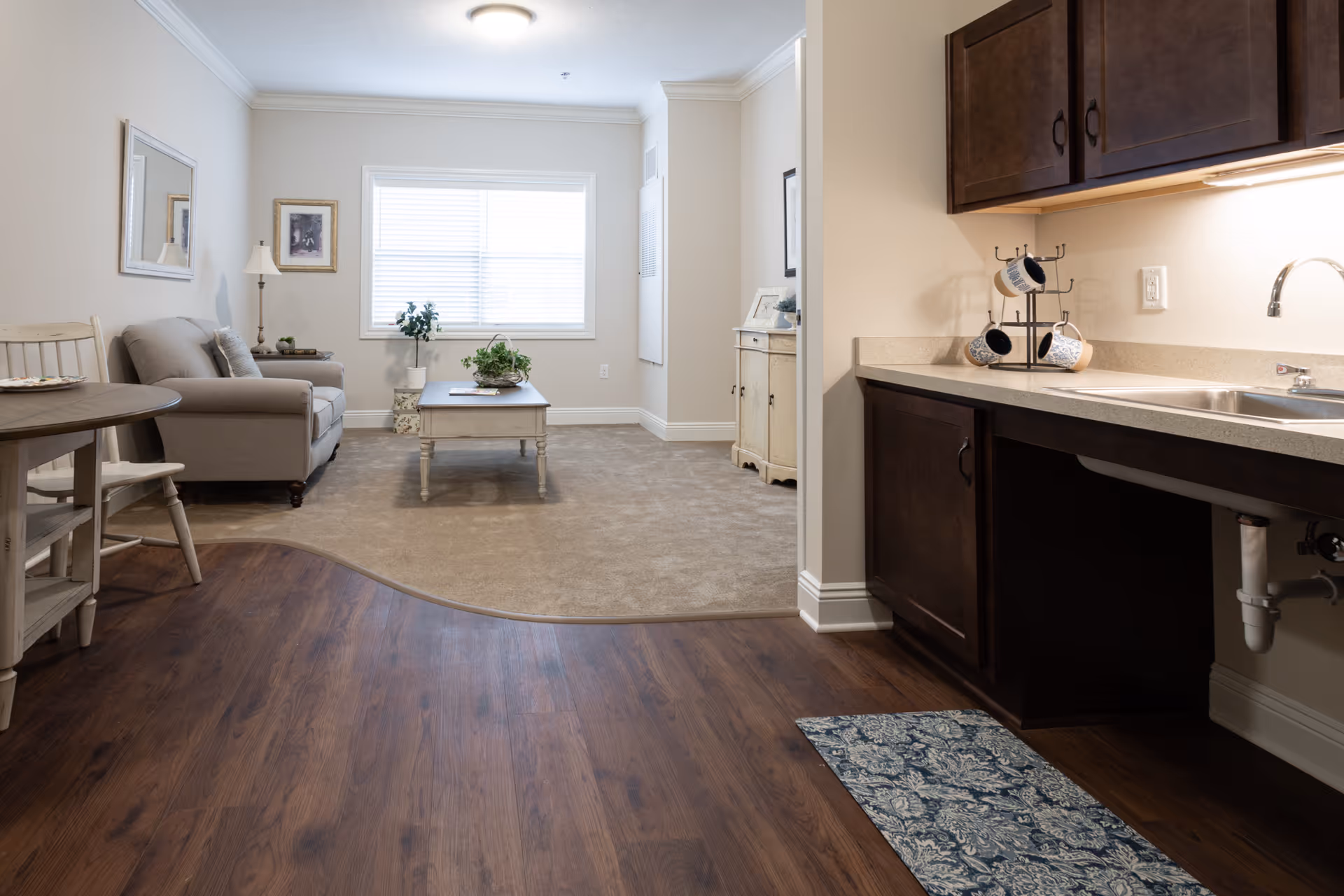 Interior view of a senior living facility showing a small living room area with a beige sofa, a coffee table with a plant, a window with blinds, and a side table with a lamp. Adjacent to the living room is a kitchenette with dark wood cabinets, a countertop, a sink, and a mug holder with mugs. The floor transitions from wood in the kitchenette area to carpet in the living room.