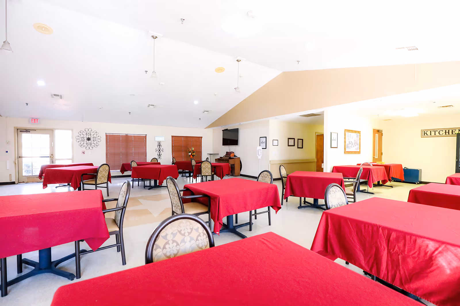 A bright dining room with multiple tables covered in red tablecloths and chairs with patterned upholstery. The room has beige walls, a high ceiling, and windows with closed blinds. There is a door on the left side and a sign labeled 'KITCHEN' on the far right wall.