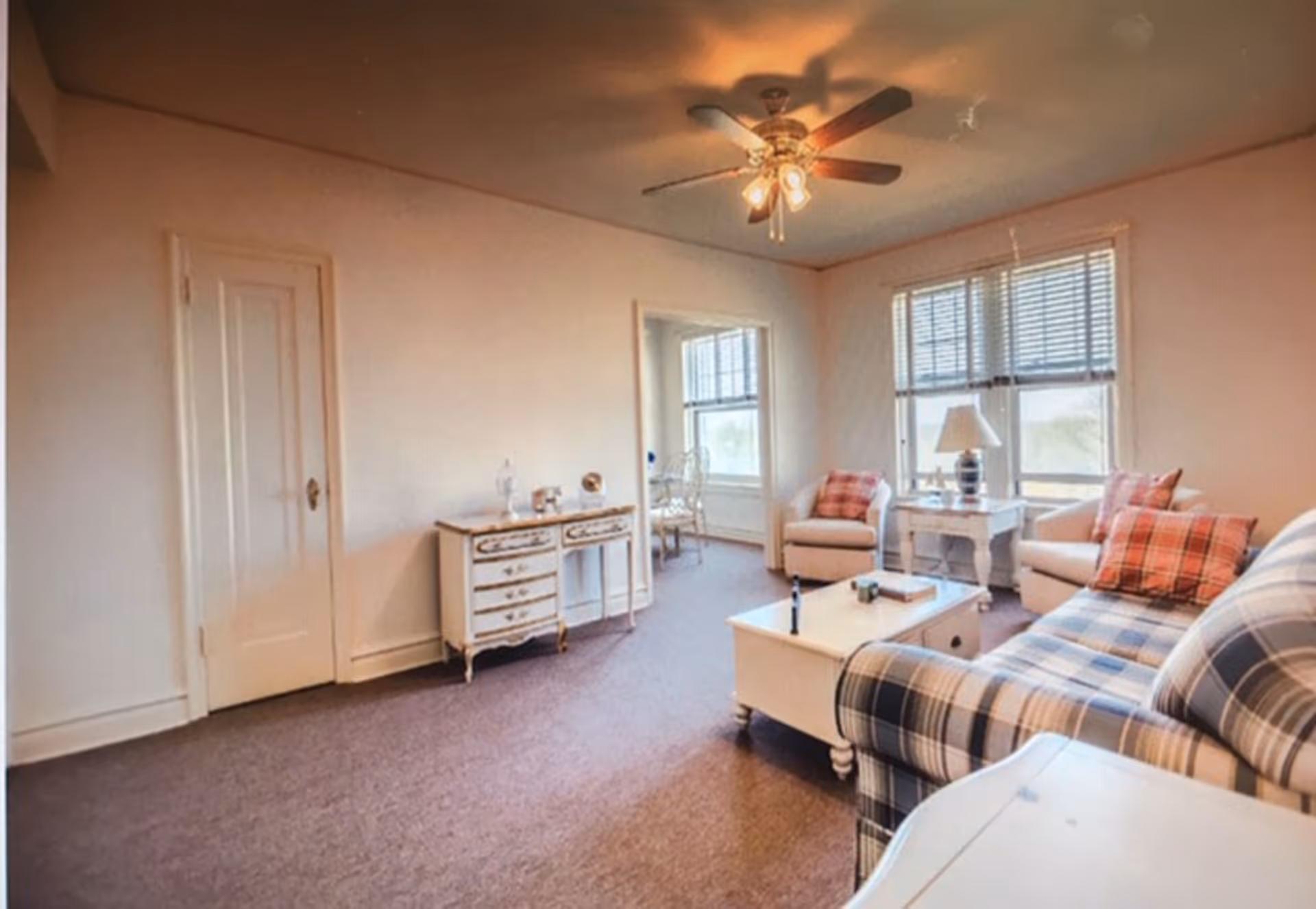 A cozy living room with a plaid sofa, two armchairs with red plaid pillows, a white coffee table, a side table with a lamp, and a ceiling fan with lights. There is a small white dresser against the wall and a window with blinds letting in natural light.