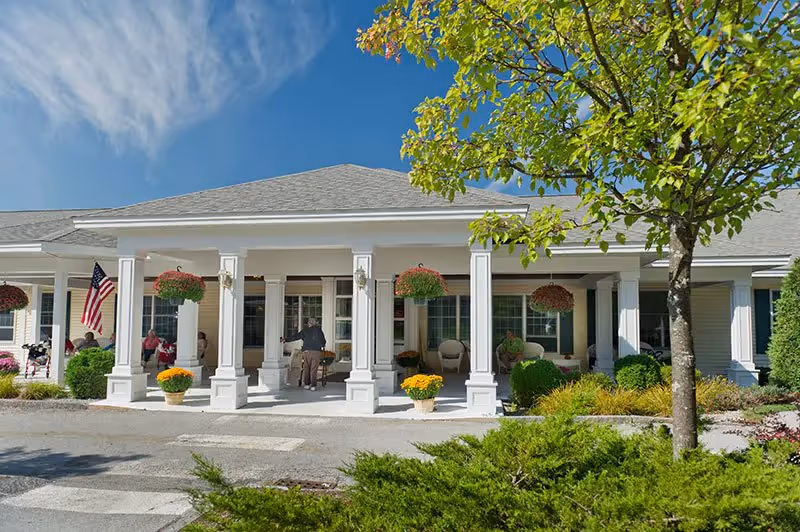 Front exterior view of a senior living facility with a covered porch supported by white columns. Several people are sitting and standing on the porch, which is decorated with hanging flower baskets and potted plants. There is a tree and shrubbery in the foreground under a clear blue sky.