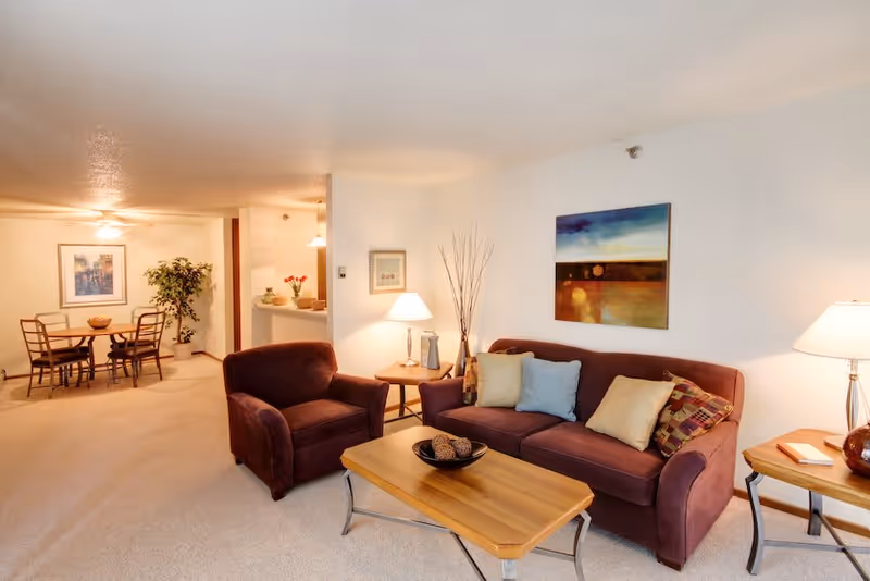 A cozy living room area with a maroon sofa and matching armchair, a wooden coffee table with decorative items, two side tables with lamps, and a colorful abstract painting on the wall. In the background, there is a dining area with a round table and four chairs, a potted plant, and a framed picture on the wall.