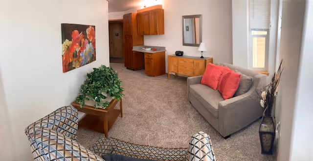A cozy living room area with a gray sofa adorned with red pillows, two patterned armchairs, a wooden coffee table with a green plant, a wooden dresser with a lamp and a mirror above it, and a colorful floral painting on the wall. The room has beige carpet and white walls, with a window letting in natural light.