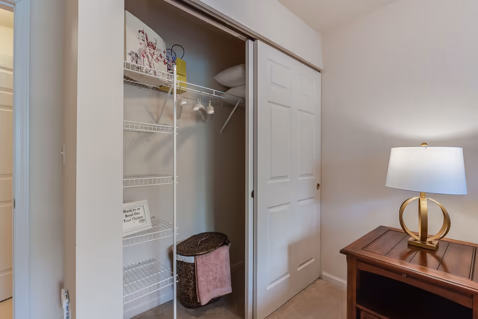 Open closet with white wire shelving and a laundry hamper beside a wooden nightstand topped by a gold lamp.