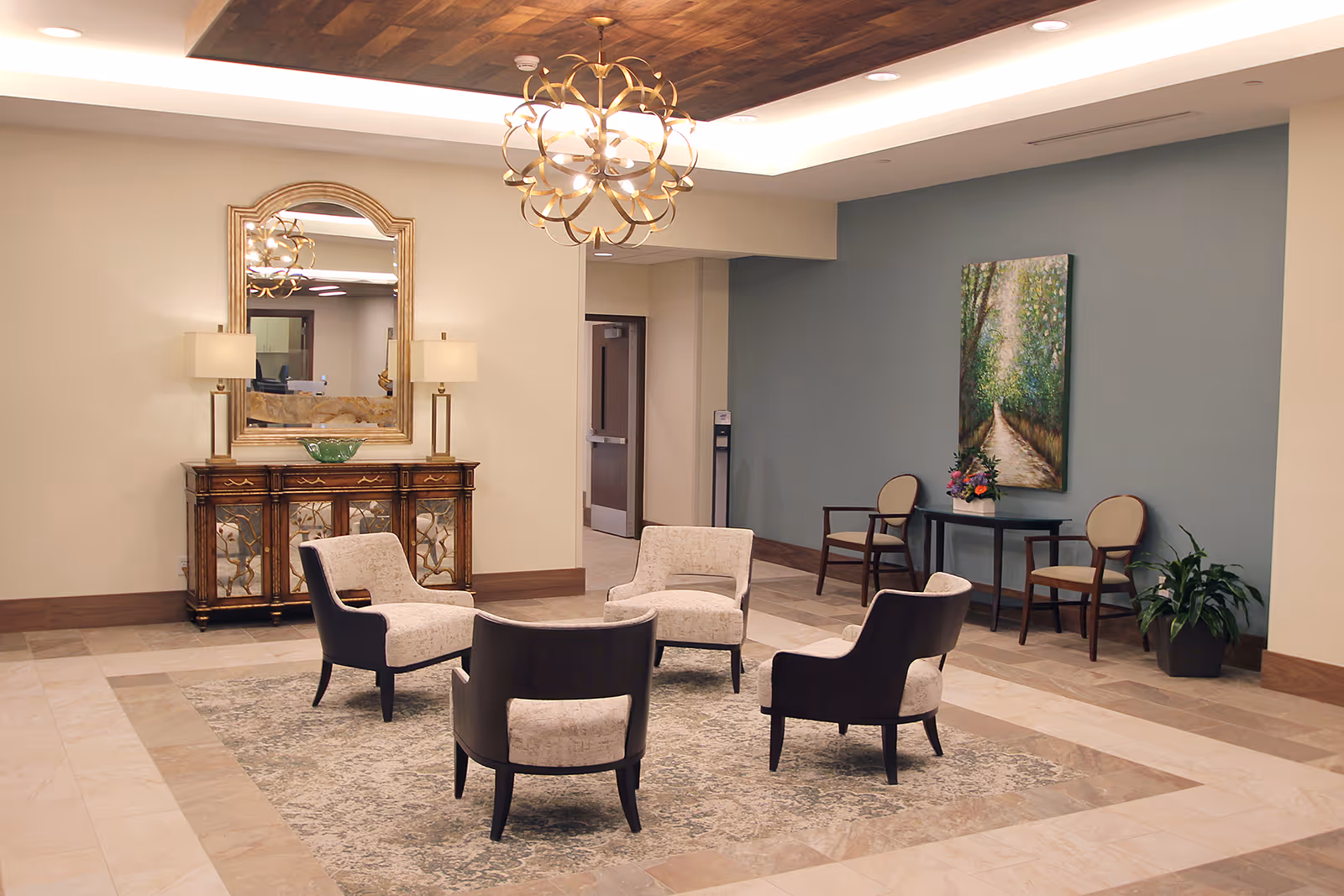 Bright, elegant lobby seating area with four upholstered chairs arranged on a rug, a mirrored console, artwork, and a chandelier.