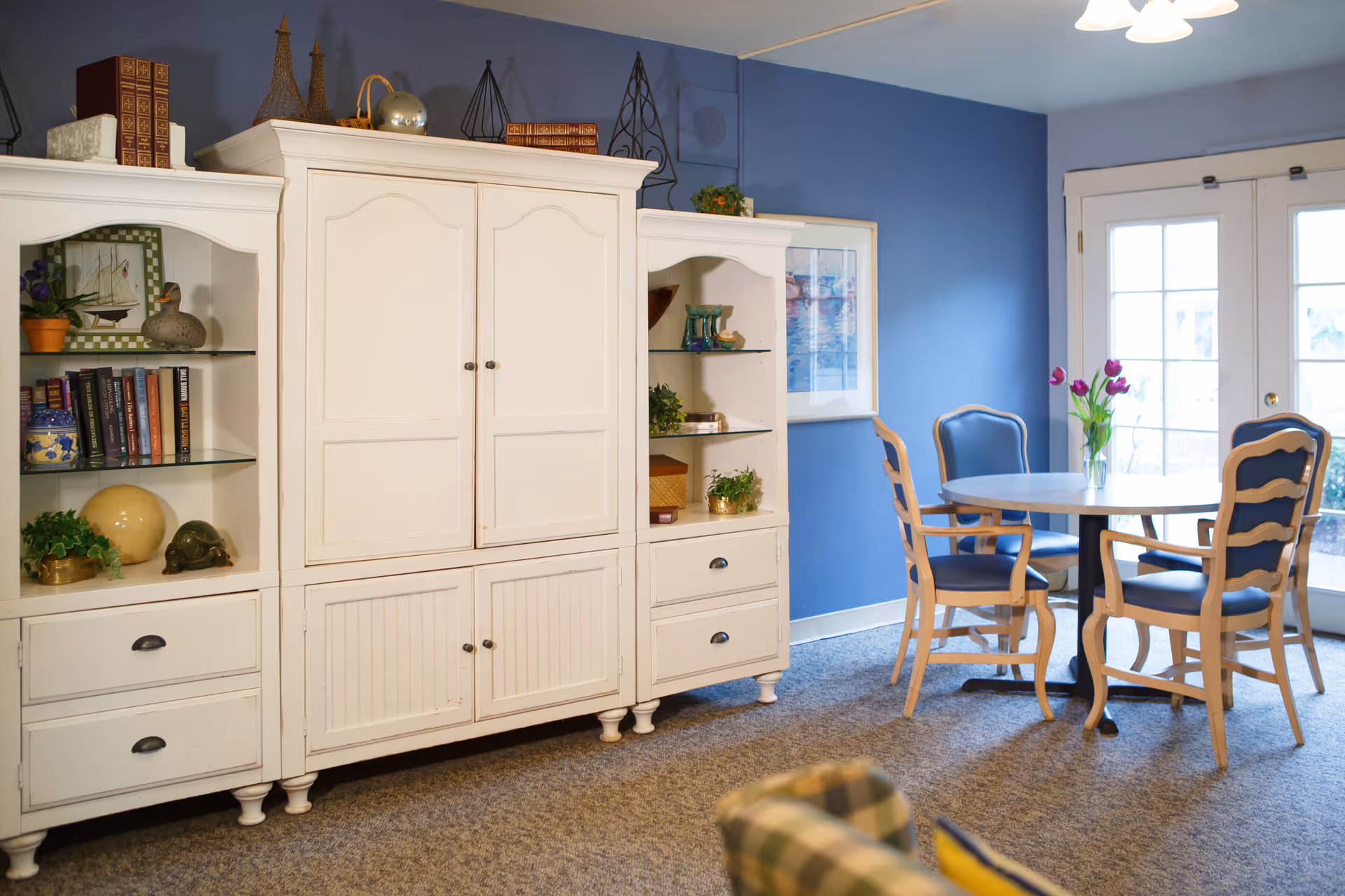 A cozy interior room with a white cabinet filled with books, decorative items, and plants on the left side. To the right, there is a round table with four wooden chairs featuring blue cushions. A vase with purple tulips is placed on the table. The walls are painted blue, and there are glass doors letting in natural light.