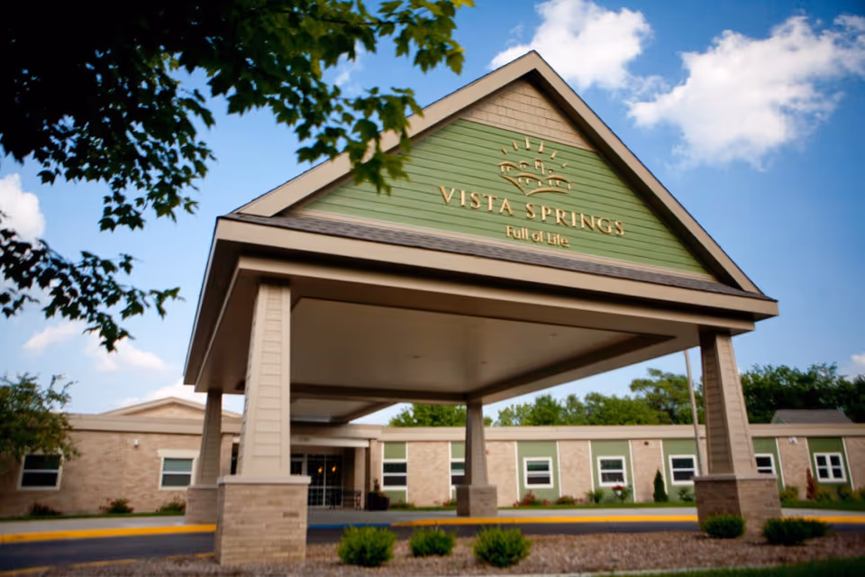 Exterior view of a senior living facility entrance with a covered drop-off area supported by columns. The building has a sign that reads 'Vista Springs Full of Life' on a green panel above the entrance. Trees and shrubs surround the building under a partly cloudy sky.