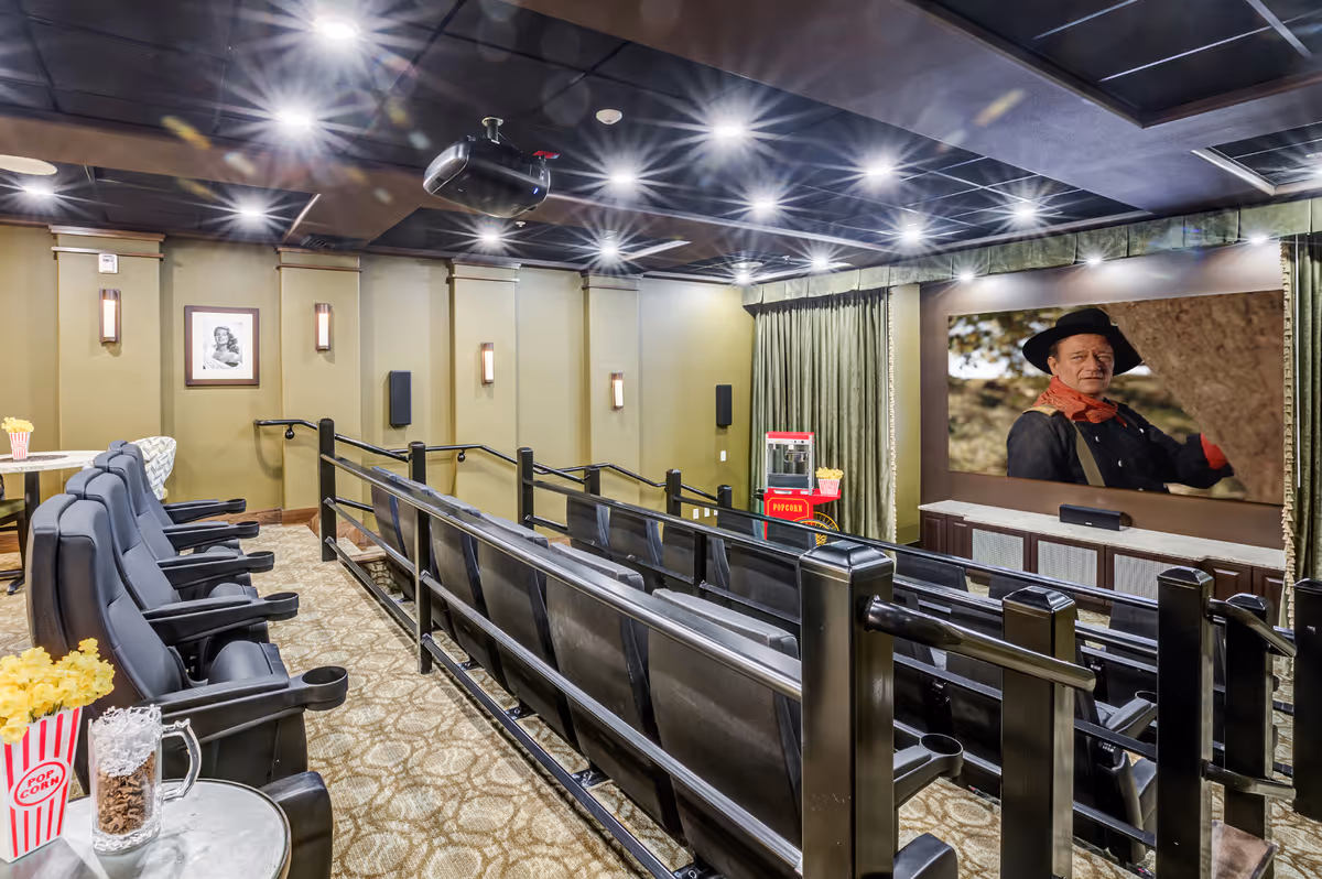 Interior of a small theater room with two rows of black reclining seats facing a large screen showing a man in a cowboy hat. The room has carpeted floors, green curtains, wall sconces, and a popcorn machine in the corner. A small table with popcorn and a drink is visible in the foreground.