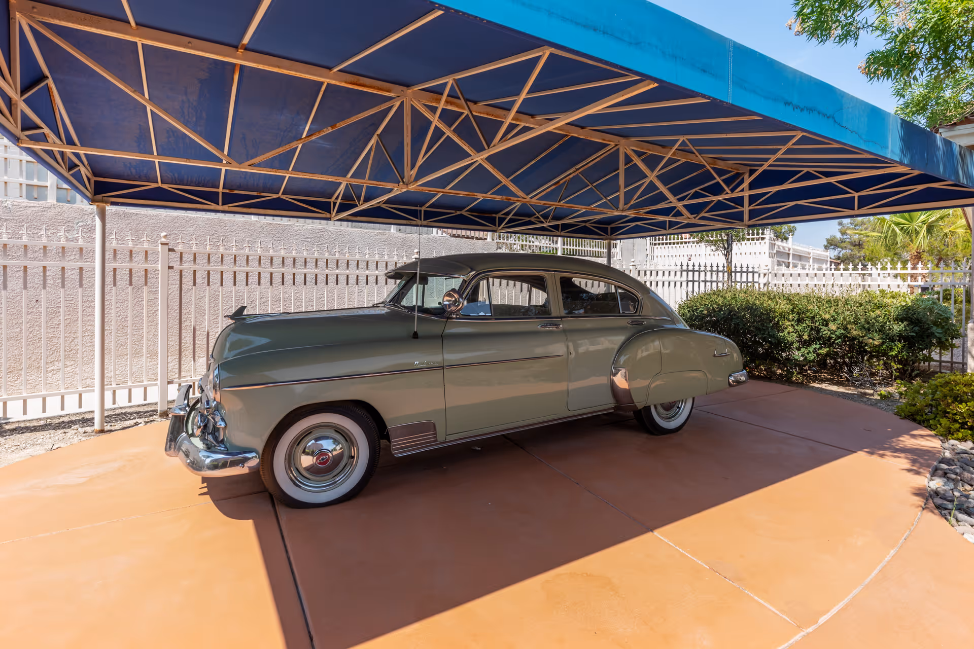 A vintage green car parked under a blue canopy in an outdoor area with a white fence and greenery in the background.