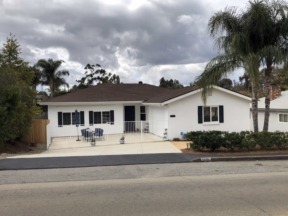 Single-story white ranch-style house with a gated front patio, outdoor seating, palm trees, and a cloudy sky.
