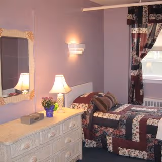 Small bedroom with a twin bed in a patchwork quilt, a dresser with a lamp and mirror, and lavender walls.