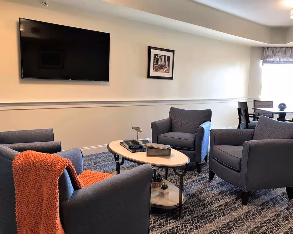 Cozy seating area with four gray armchairs arranged around an oval coffee table, a wall-mounted TV, and a small dining table by a window.