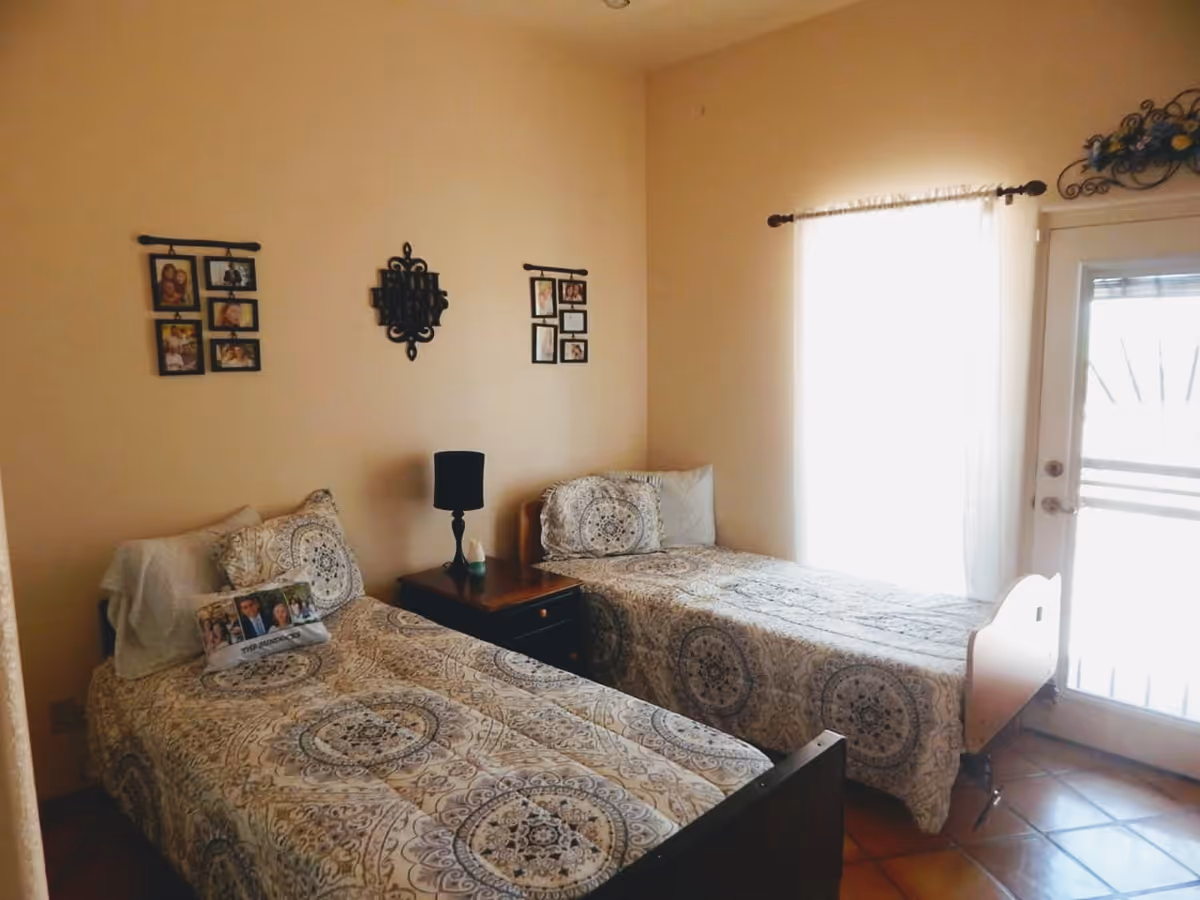 A bedroom with two single beds placed parallel to each other, each with patterned bedspreads and pillows. Between the beds is a wooden nightstand with a black lamp and a small decorative item. The walls are beige and decorated with framed photos and a decorative wall piece. A window with sheer white curtains and a glass door letting in natural light are visible on the right side.