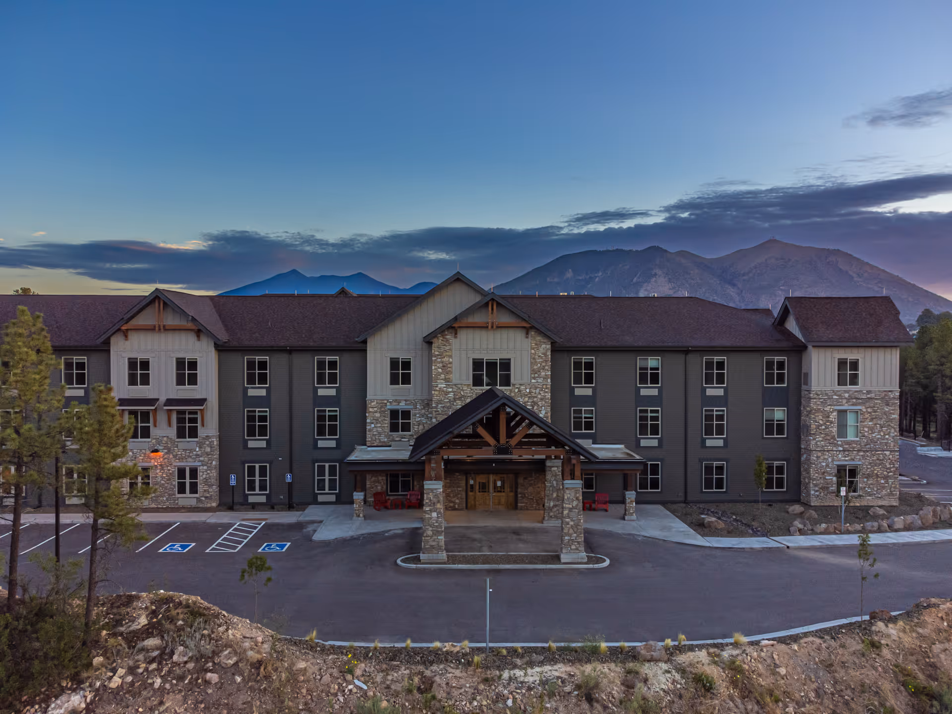 Front exterior of a three-story senior living building with a covered entrance and mountains in the background at dusk.