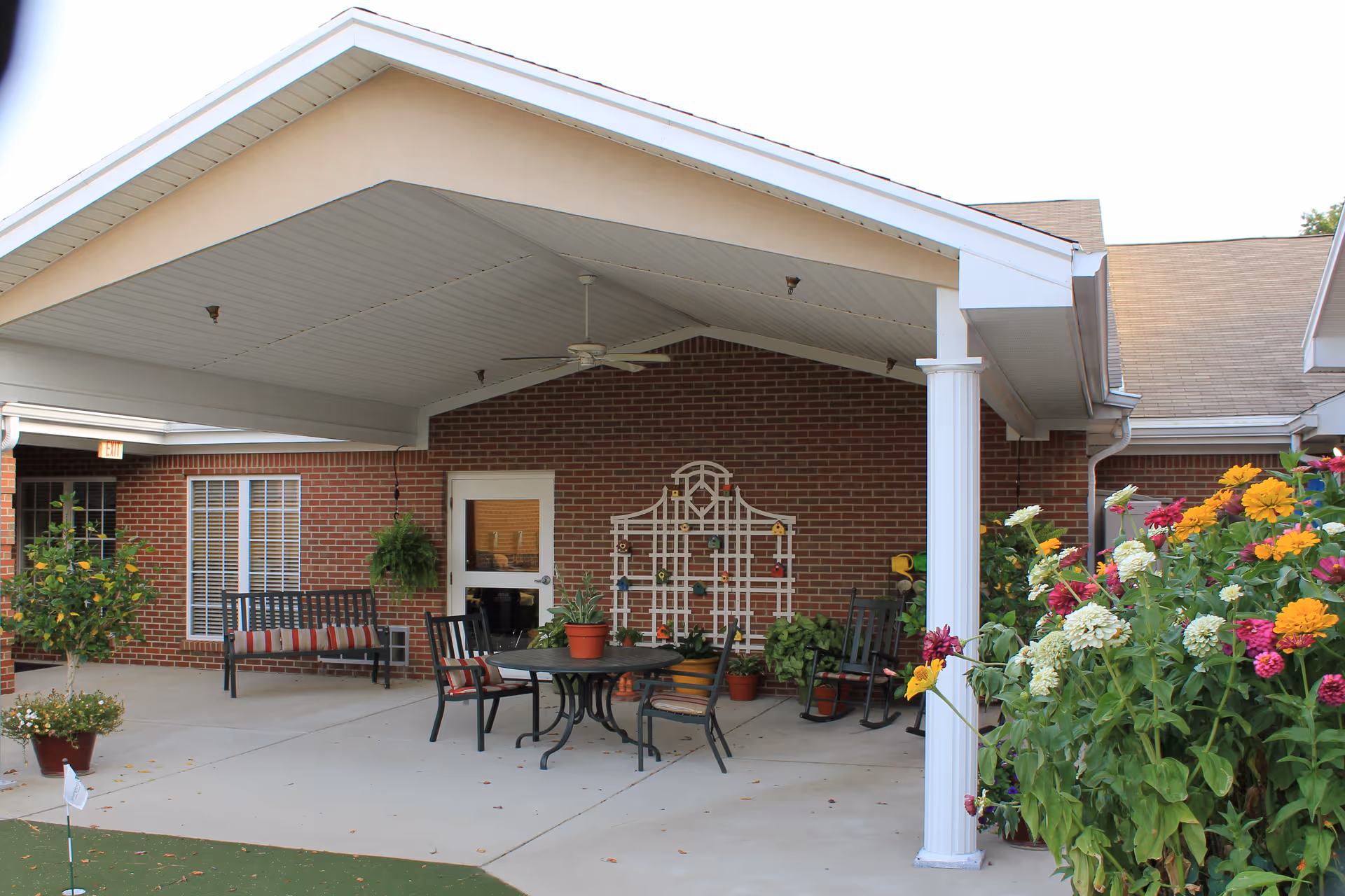 Covered outdoor patio area with a round table and chairs, a bench with cushions, rocking chairs, potted plants, and colorful flowers in the foreground. The patio is attached to a brick building with a white door and windows.