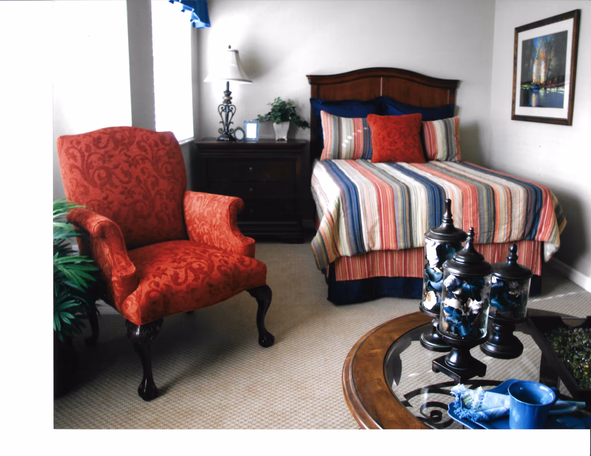 A bedroom with a wooden bed featuring a striped bedspread in blue, red, and beige tones, and multiple pillows including a red decorative one. To the left of the bed is a dark wooden nightstand with a decorative lamp, a small plant, and a picture frame. In front of the bed is a round glass-top table with three decorative jars and a blue cup with a napkin. A red upholstered armchair with dark wooden legs is positioned near a window with a blue valance. A framed picture hangs on the wall to the right of the bed.