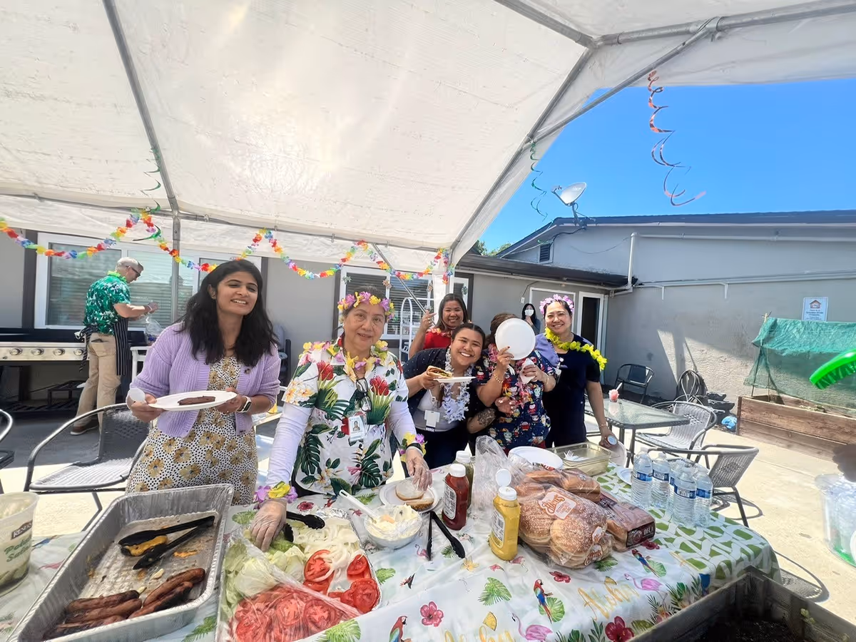 A group of six people enjoying an outdoor barbecue under a white canopy. They are smiling and holding plates with food. The table in front of them is covered with a floral tablecloth and has various food items including buns, condiments, sliced tomatoes, onions, lettuce, and water bottles. The background shows a building wall and outdoor chairs.