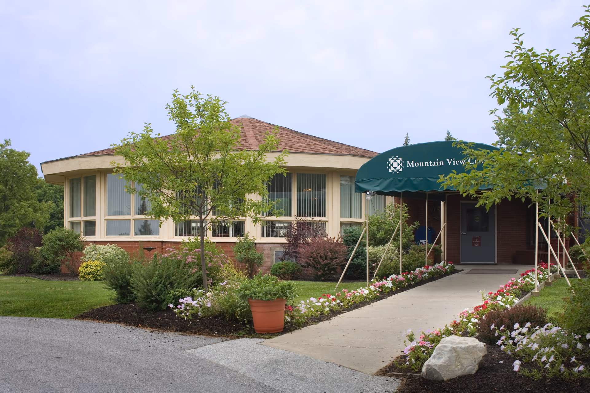 Exterior view of Mountain View Center building with a green awning over the entrance. The building has large windows and is surrounded by well-maintained landscaping including trees, shrubs, and flower beds along a paved walkway.