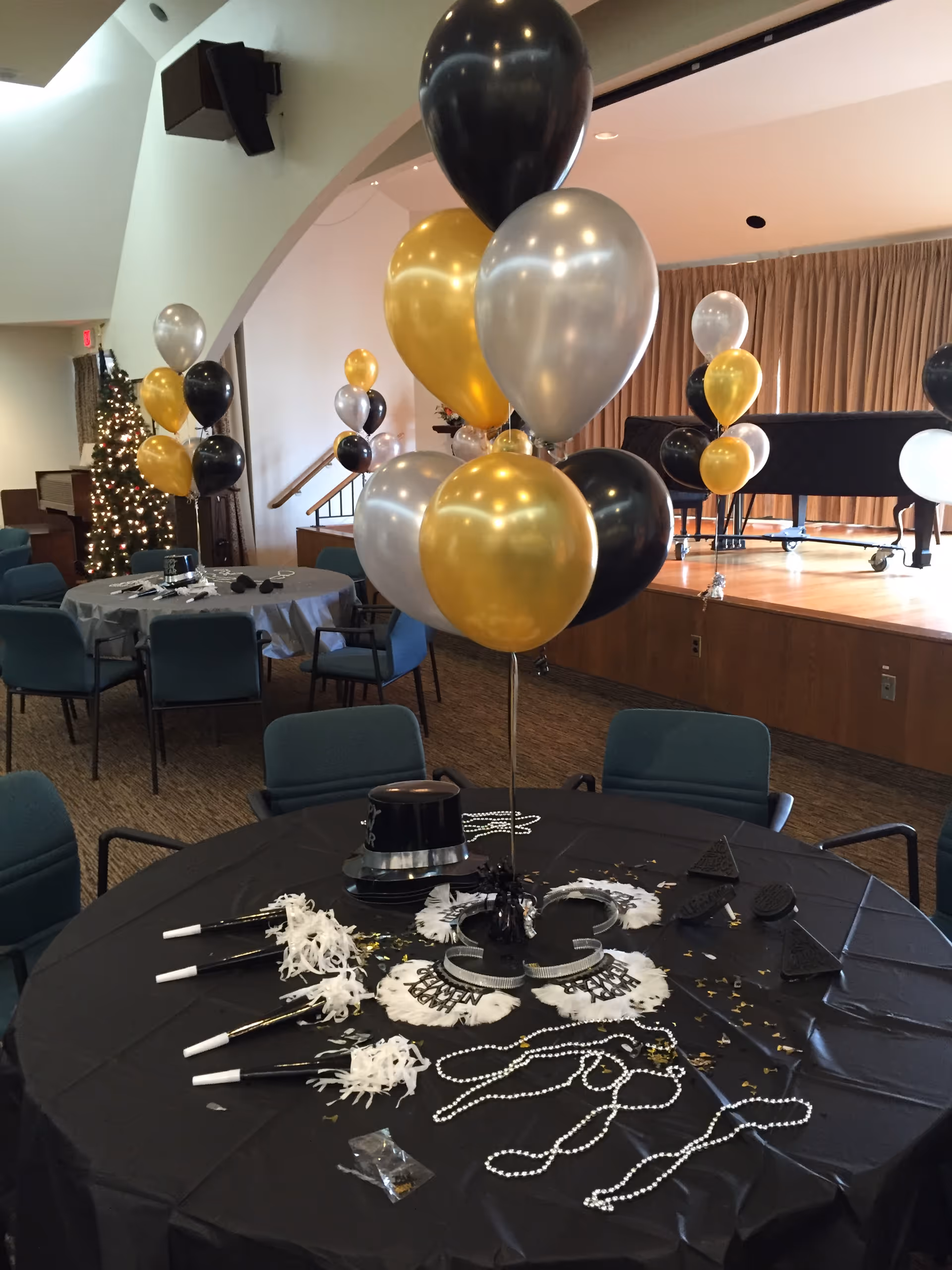 A decorated event room with round tables covered in black tablecloths, adorned with black, gold, and silver balloons. The tables have party hats, noise makers, bead necklaces, and New Year's Eve themed decorations. In the background, there is a stage with a piano and a Christmas tree with lights.