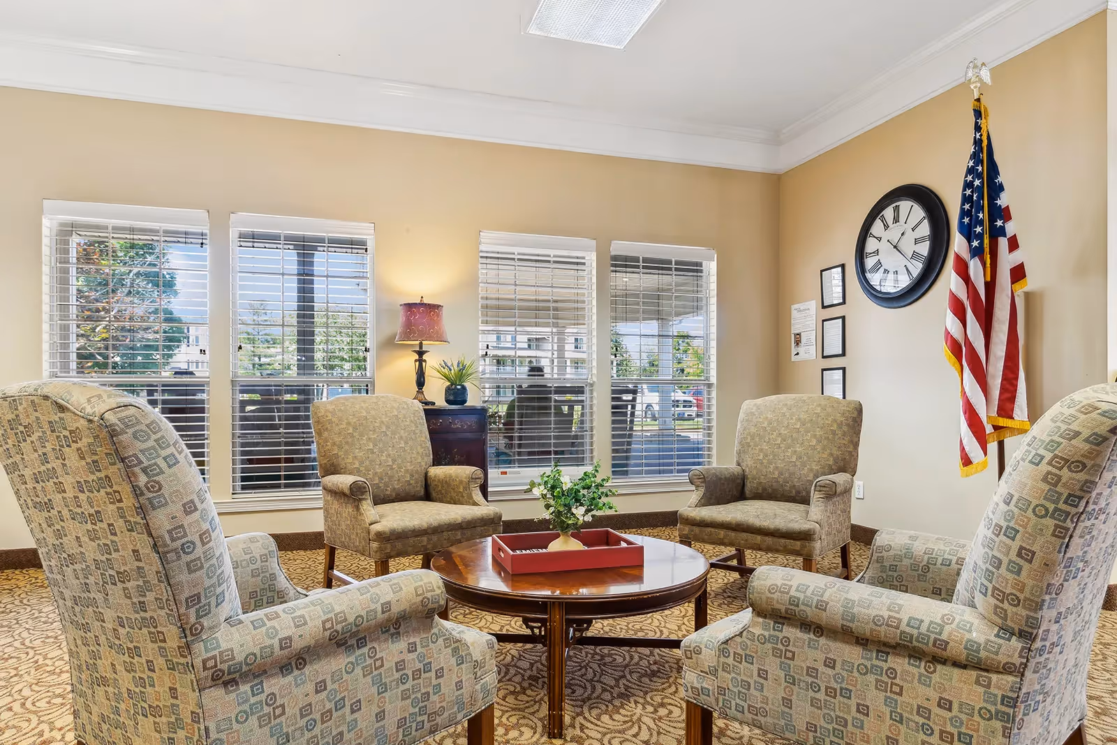 A cozy sitting area with four patterned armchairs arranged around a round wooden coffee table with a red tray and a small plant. The room has beige walls, a large clock, an American flag, and three windows with white blinds letting in natural light. A small table with a lamp and a plant is placed against the wall between the windows.