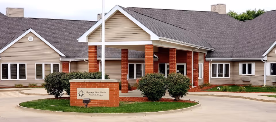 Exterior front view of Regency Care Center Assisted Living building with a covered entrance supported by brick columns, surrounded by bushes and a circular driveway.