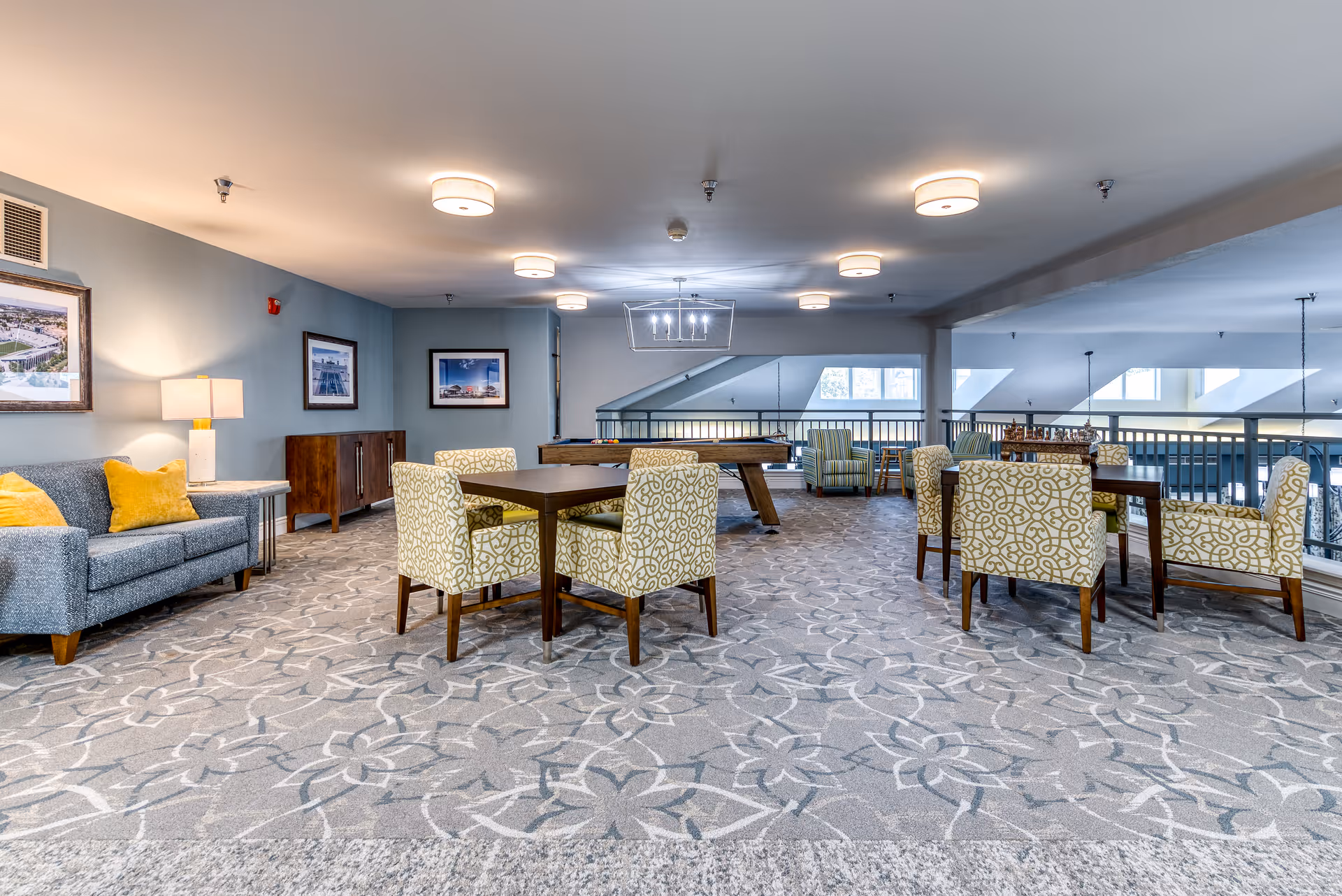A spacious senior living common area with patterned carpet, several tables surrounded by patterned chairs, a gray sofa with yellow pillows, framed pictures on the walls, and a pool table in the background under ceiling lights.