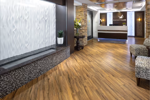 Reception lobby with a front desk bearing the 'Elevate Care' logo, decorative stone columns, seating, and a textured water feature.