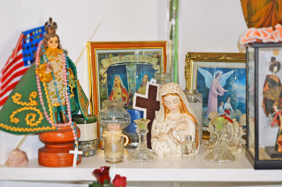 A shelf displaying various religious items including statues of the Virgin Mary and a crowned figure dressed in green, framed religious pictures, a cross, candles, and glass candle holders.