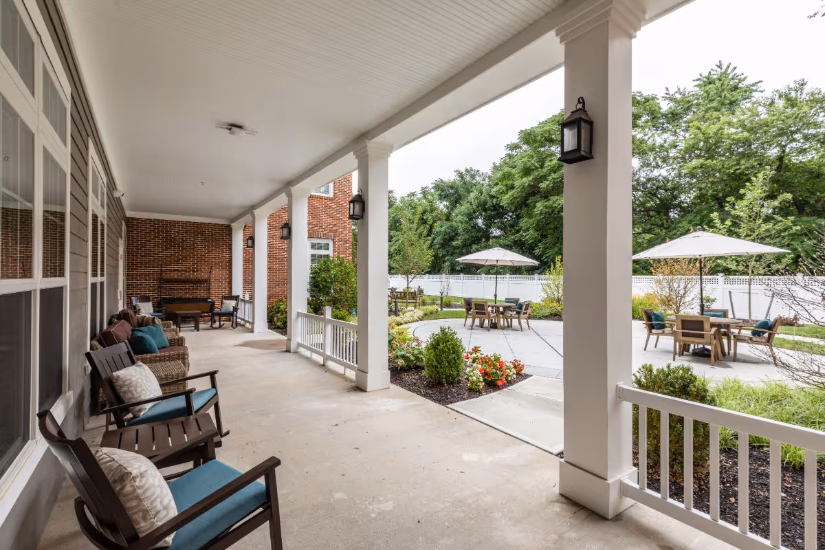 Covered porch with cushioned chairs overlooking a landscaped courtyard with tables and umbrellas.