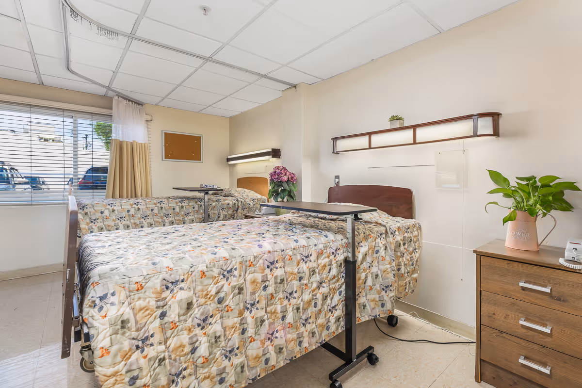 A bright healthcare facility room with two hospital beds covered in floral patterned quilts. Each bed has an overbed table, and there is a wooden nightstand with a potted plant and a telephone. A large window with blinds and curtains lets in natural light. The walls are light-colored, and there is a corkboard and a wall-mounted light fixture above one of the beds.