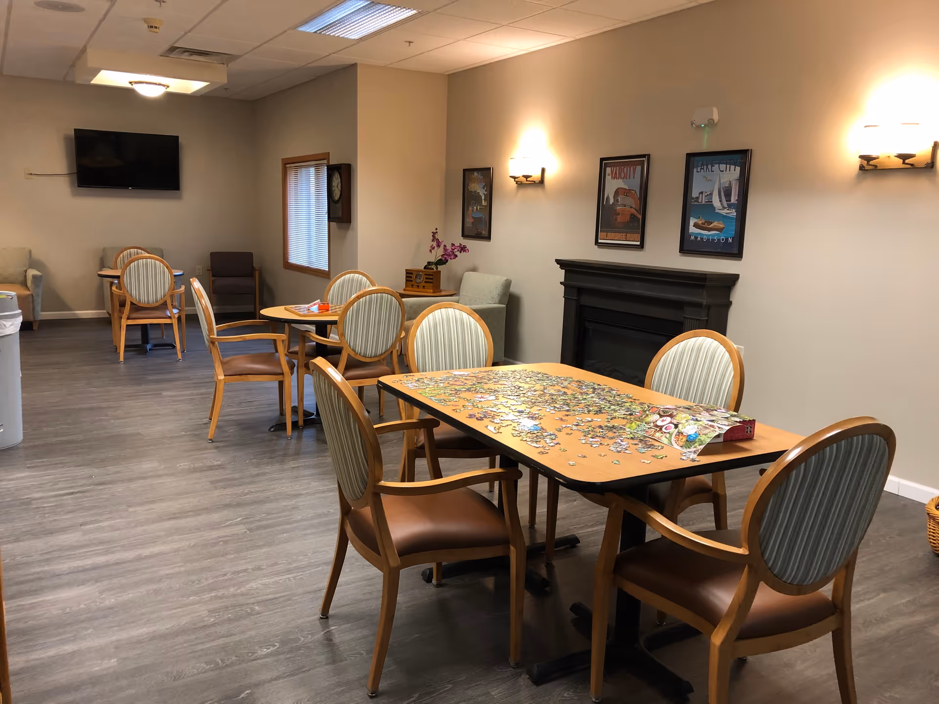A cozy common area in Coventry Village with several wooden tables and chairs arranged for social activities. One table in the foreground has a partially completed jigsaw puzzle. The room features a wall-mounted TV, framed artwork, a fireplace, and soft lighting from wall sconces. The floor is covered with wood-style flooring.
