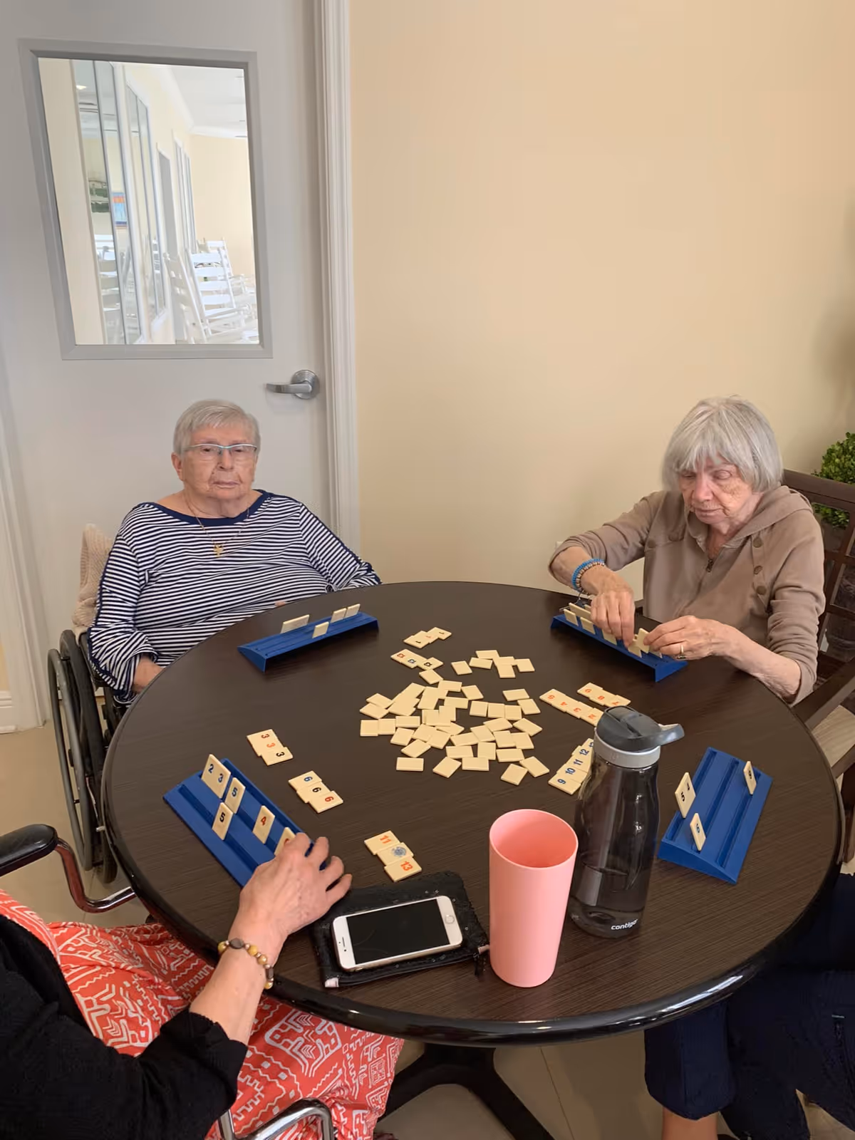 Three elderly women sitting around a round table playing a tile-based game in a room with beige walls and a white door with a window. On the table are game tiles, a pink cup, a water bottle, and a smartphone.