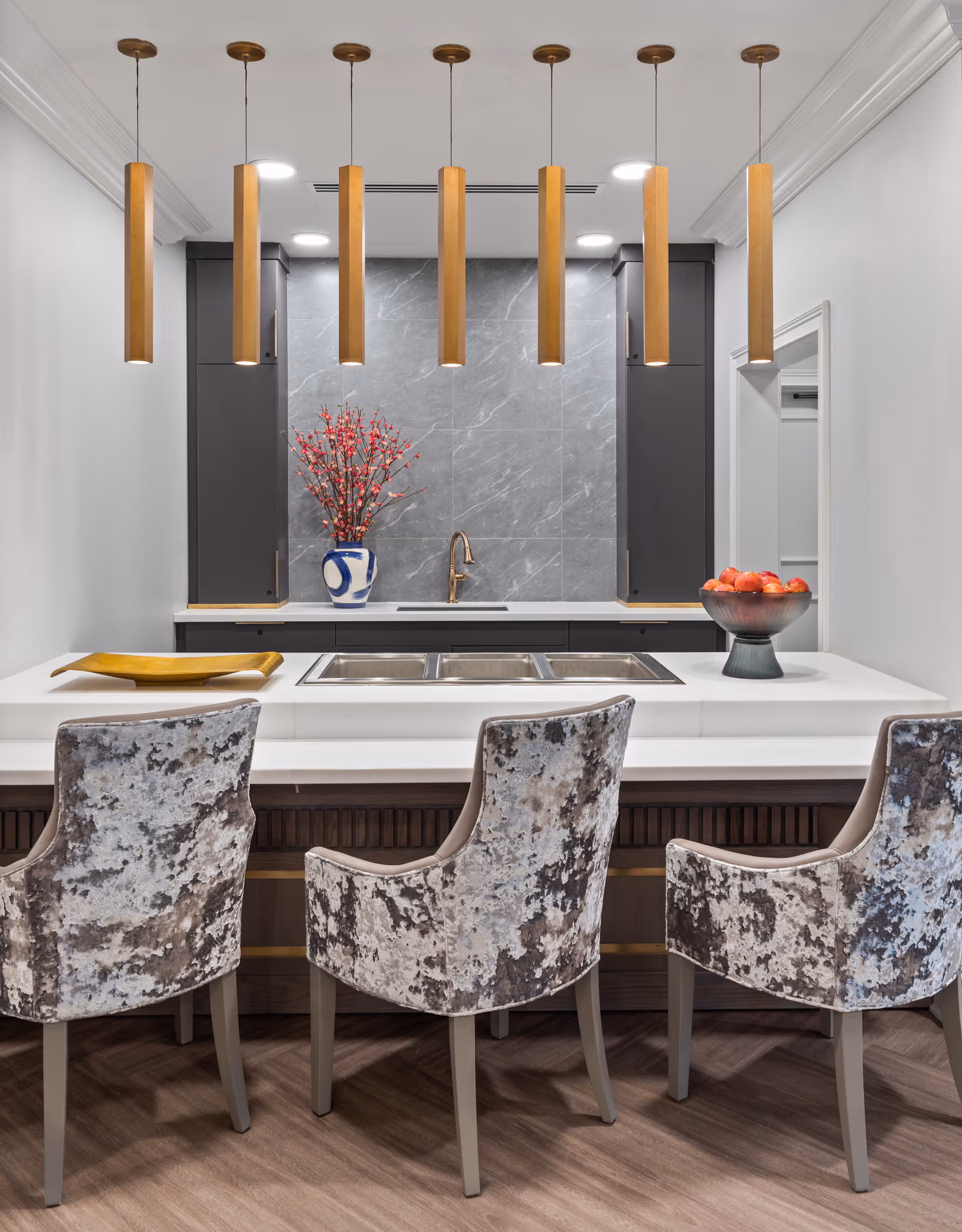 Modern kitchen area with a white countertop island featuring three patterned upholstered chairs. Above the island hang seven vertical wooden pendant lights. The back wall has a gray marble backsplash with a gold faucet and a blue and white vase with red flowers. A bowl of red apples and a gold tray are placed on the countertop.