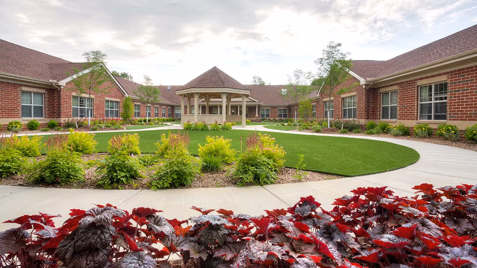 Outdoor courtyard area of Pomeroy Living Rochester Assisted Living and Memory Care featuring a central gazebo surrounded by a circular walkway, green lawn, and landscaped plants with red and green foliage. The courtyard is enclosed by single-story brick buildings with multiple windows under a cloudy sky.