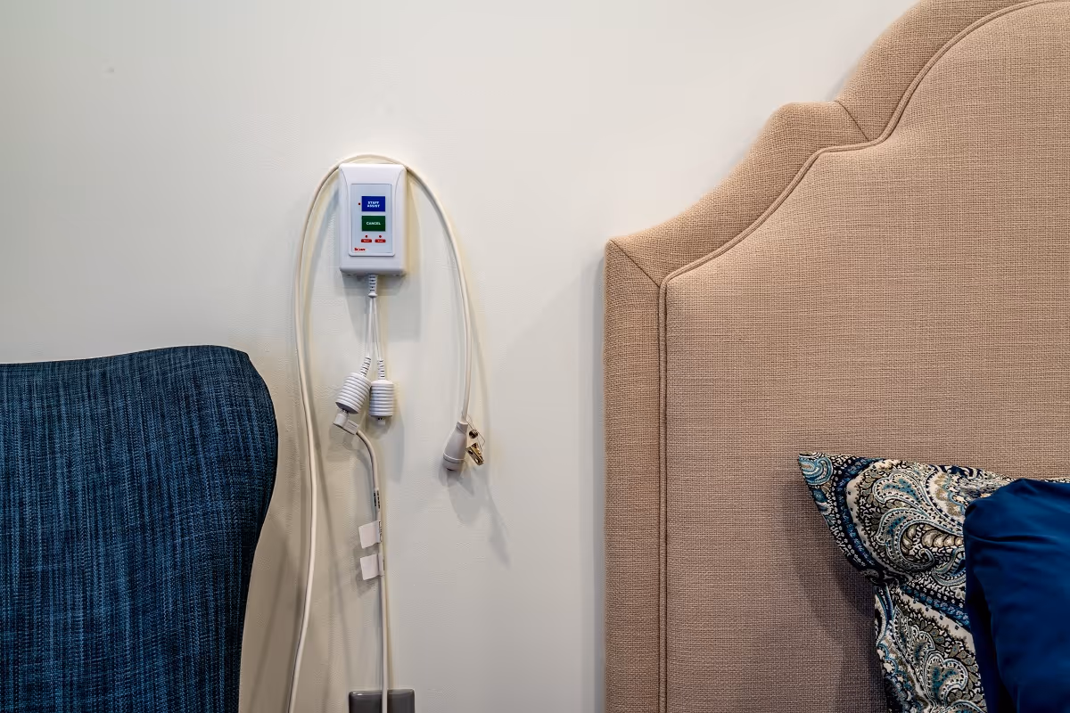 Close-up view of a beige upholstered headboard with a patterned pillow and a blue pillow on a bed. Next to the headboard is a blue upholstered chair and a wall-mounted emergency call button with cords hanging down.