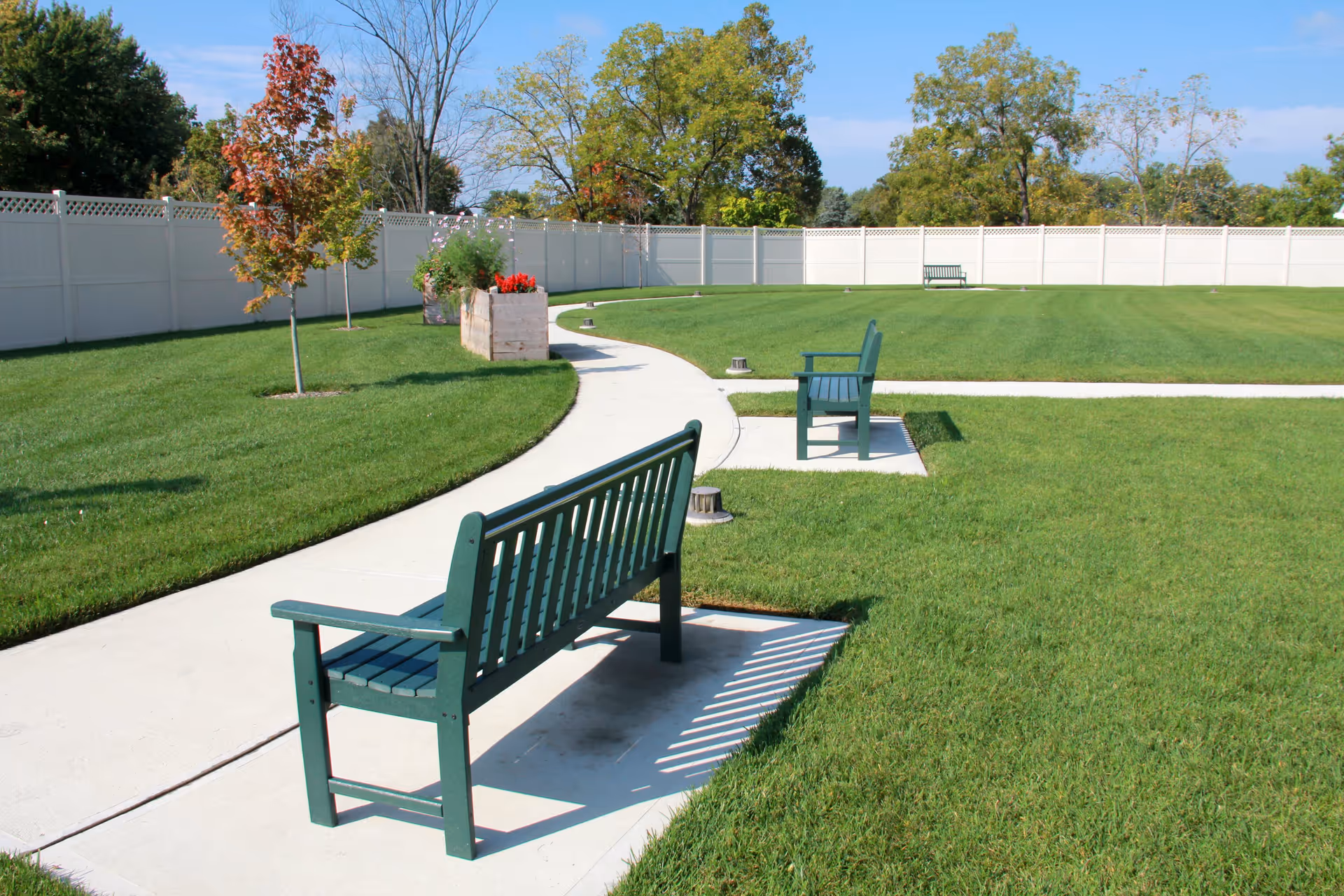 A well-maintained outdoor garden area with green grass, a curved concrete walkway, and several green benches placed along the path. There are small trees and flower planters near a white privacy fence, with a clear blue sky and some trees in the background.