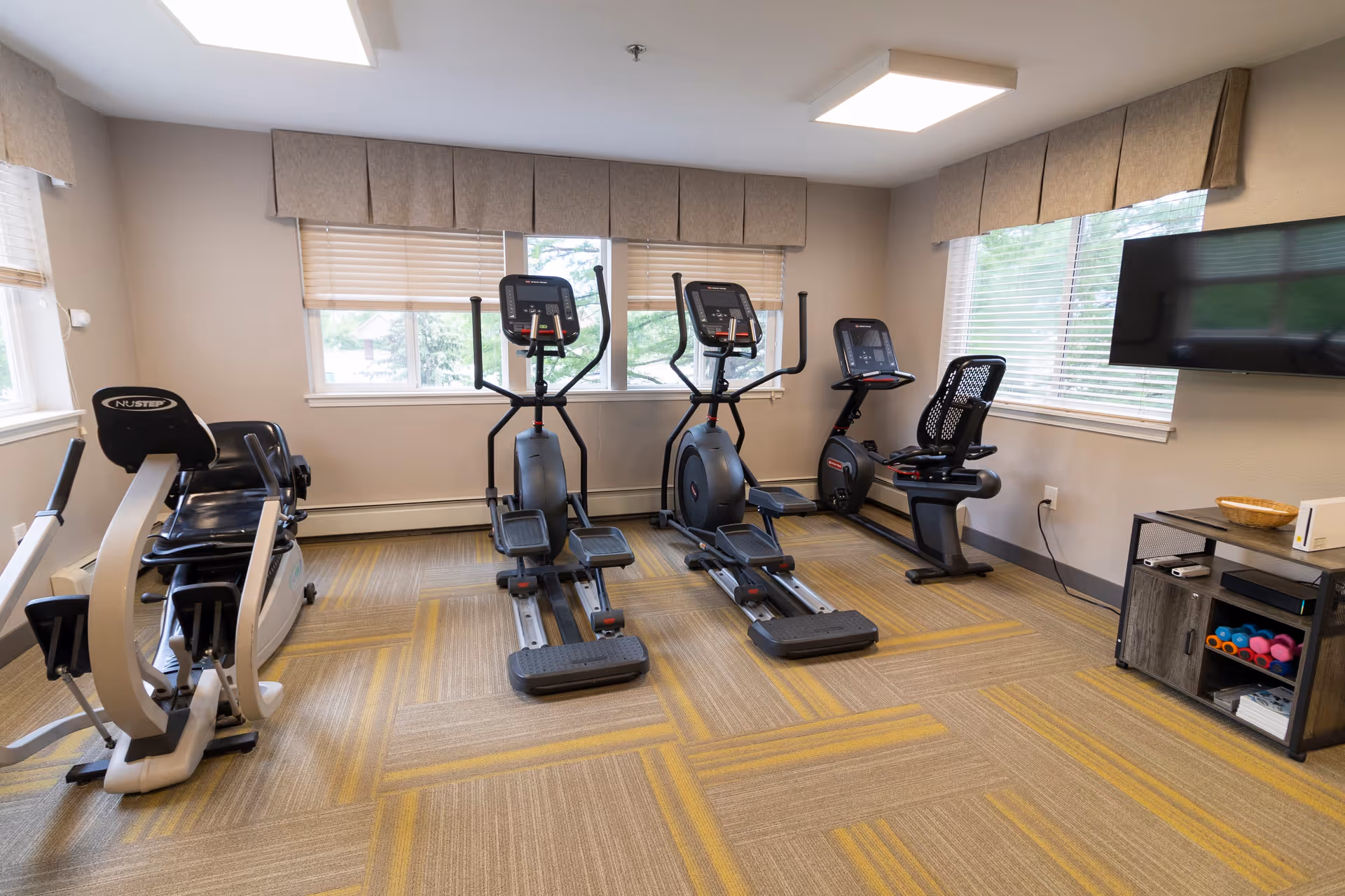 A small fitness room with exercise equipment including a recumbent bike, two elliptical machines, and a stationary bike. The room has beige walls, large windows with blinds and valances, a wall-mounted TV, and a small cabinet holding colorful dumbbells and other items.