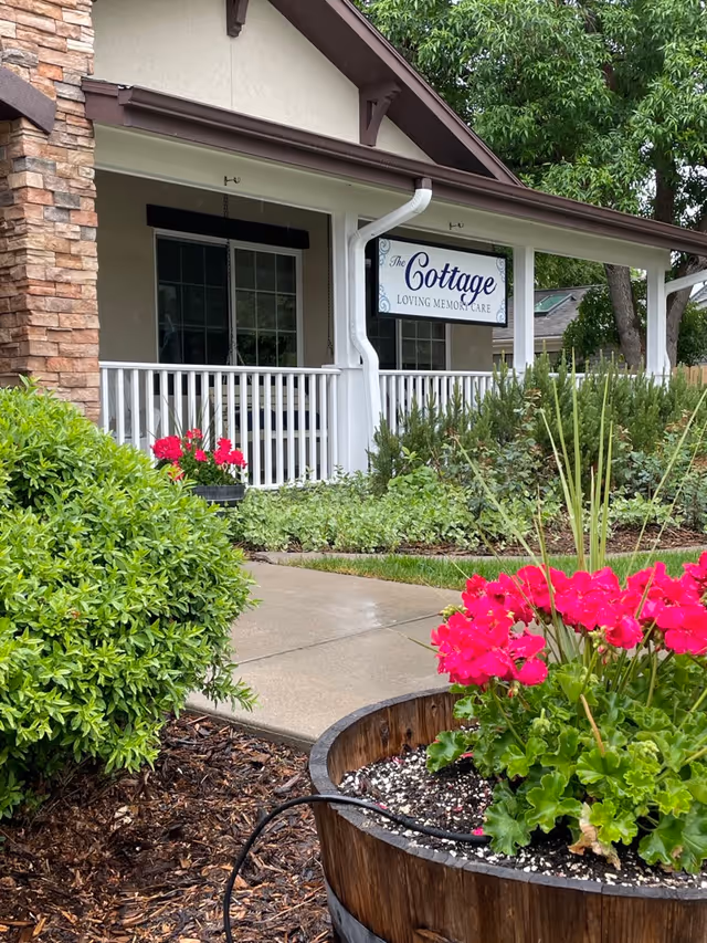Front porch of a building with a white railing and a sign that reads 'The Cottage Loving Memory Care'. There are green bushes and a wooden planter with bright pink flowers in the foreground, along with a concrete walkway leading to the porch.