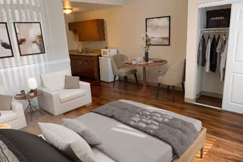 Studio-style interior with a bed in the foreground, a seating area and small dining table, kitchenette cabinets, and an open closet.