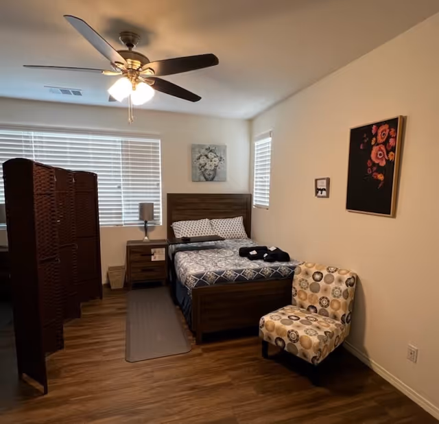 A cozy bedroom with a wooden bed frame and a patterned bedspread. There are two pillows on the bed and folded towels placed on top. Next to the bed is a wooden nightstand with a lamp. A decorative folding screen is positioned near the window with closed blinds. The room has wood flooring, a ceiling fan with lights, and two framed floral artworks on the walls. A patterned upholstered chair is placed against one wall.